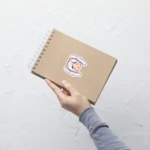 Fire Emblem sticker of Female Robin (small, 3x3) applied to a brown spiral notebook held in one hand.