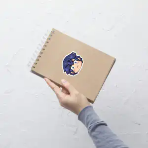 Fire Emblem sticker of Chrom (medium, 4x4) centered on a tan notebook against a white textured wall.