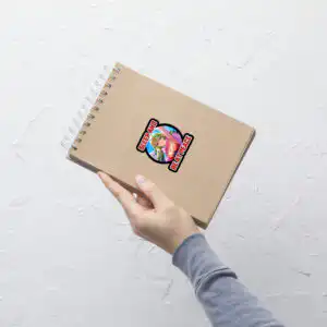 Small Fire Emblem Sticker featuring Adult Tiki applied to a brown spiral notebook, held in one hand.